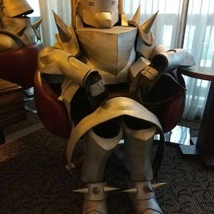 Peut inclure: Un costume de robot argenté et doré avec un casque pointu et une armure à pointes. Le robot est assis sur une chaise rouge, les bras croisés.