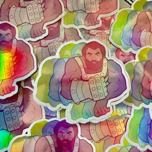 May include: A stack of iridescent stickers featuring a cartoon illustration of a muscular, bearded man with a serious expression. The man is wearing a metal arm brace and is flexing his bicep.