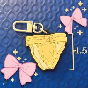 May include: A yellow and black keychain charm shaped like a diaper, measuring 1.5 inches. It has a white clasp and ring. Pink bows and yellow stars are drawn around the charm on a blue background.