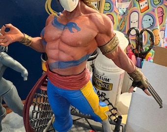 May include: A detailed figurine of a muscular character wearing a white mask with a 'J' on it, blue and yellow pants, and a red sash. The figure has long blonde hair and is posed with a weapon. The figurine is on a gray base.