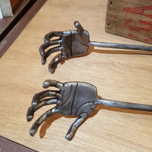 Puede incluir: Dos pinzas de metal en forma de mano con un acabado rústico. Las pinzas están hechas de metal y tienen un diseño en forma de mano. Las pinces están sobre una superficie de madera.