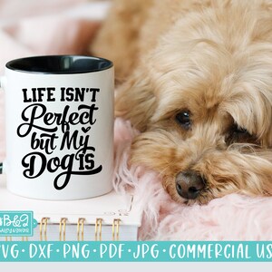 Puede incluir: Una taza de cerámica blanca con interior y asa negros. La taza muestra el texto "LIFE ISN'T Perfect but My Dog is" en escritura negra. Un perro esponjoso, de color marrón claro, descansa sobre una superficie rosa junto a la taza.