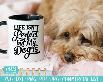 May include: A white ceramic mug with a black interior and handle. The mug features the text "LIFE ISN'T Perfect but My Dog is" in black script. A fluffy, light brown dog is resting on a pink surface next to the mug.