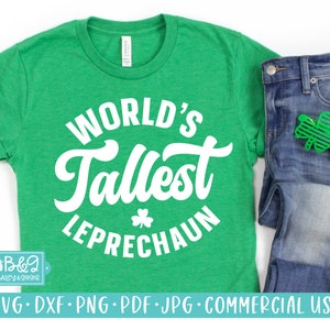 May include: A green t-shirt with white text that reads "WORLD'S Tallest LEPRECHAUN". The shirt is on a white background with a pair of blue jeans and a green shamrock.