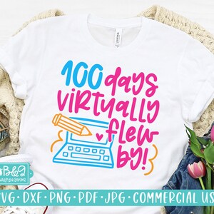 May include: A white t-shirt with the text "100 days virtually flew by!" in pink, blue, and orange. The text is surrounded by a cartoon image of a laptop with a pencil hovering over it.
