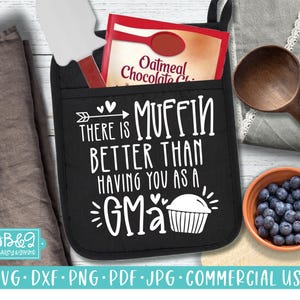 May include: Black oven mitt with the text "THERE IS MUFFIN BETTER THAN HAVING YOU AS A GMA" in white. A packet of oatmeal chocolate chip mix and a spatula are in the pocket. A wooden spoon and a bowl of blueberries are also present.