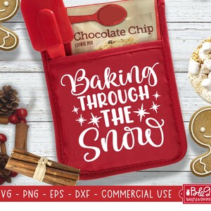 May include: Red oven mitt with the text "Baking Through The Snow" in white. The oven mitt is folded and has a pocket on the front. The pocket contains a packet of chocolate chip cookie mix.