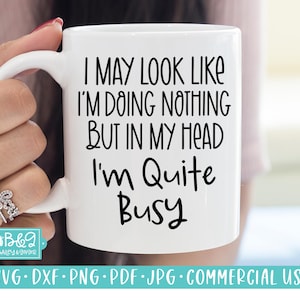 Puede incluir: Taza de cerámica blanca con el texto negro: "I MAY LOOK LIKE I'M DOING NOTHING BUT IN MY HEAD I'm Quite Busy". La taza está sujeta por una persona. La parte inferior de la taza tiene un logotipo y las palabras "SVG.DXF.PNG.PDF.JPG.COMMERCIAL USE".