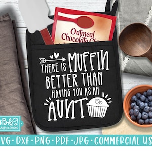 Może przedstawiać: Czarna rękawica kuchenna z białym napisem „THERE IS MUFFIN BETTER THAN HAVING YOU AS AN AUNT”. W kieszeni znajduje się paczka mieszanki płatków owsianych z kawałkami czekolady i szpatułka. Widoczna jest również drewniana łyżka i miska z jagodami.