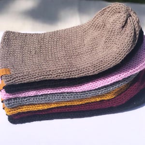 May include: A stack of six knitted beanies in various colours, including black, burgundy, mustard yellow, grey, pink, and brown. The beanies are made of a soft, textured yarn.