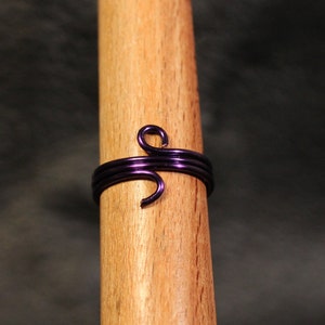 May include: A handcrafted, dark purple wire ring with a swirling design. The ring is wrapped around a wooden dowel, showcasing its shape and detail. The ring's metallic sheen contrasts with the natural wood grain.