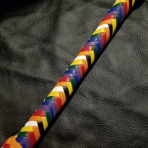 May include: A multi-colored leather whip with a braided design. The whip is made of different colored leather strips, including red, orange, yellow, green, blue, purple, and black. The whip has a rounded end.