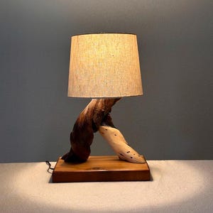May include: A wooden table lamp with a natural wood base and a beige fabric lampshade. The base is shaped like a twisted branch.