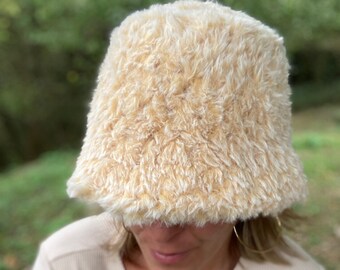 Puede incluir: Un gorro de cubo de color crema esponjoso con una textura suave y peluda.