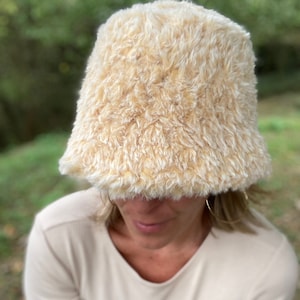 Puede incluir: Un gorro de cubo de color crema esponjoso con una textura suave y peluda.