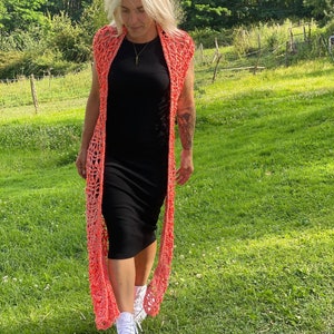 Pu&ograve; includere: Una donna indossa un cardigan di uncinetto corallo sopra un vestito nero e scarpe da ginnastica bianche. Sta camminando in un campo erboso.