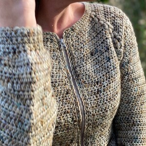 May include: A close-up of a beige crocheted cardigan with a zip fastening. The cardigan features a textured stitch pattern and a braided detail on the shoulder.