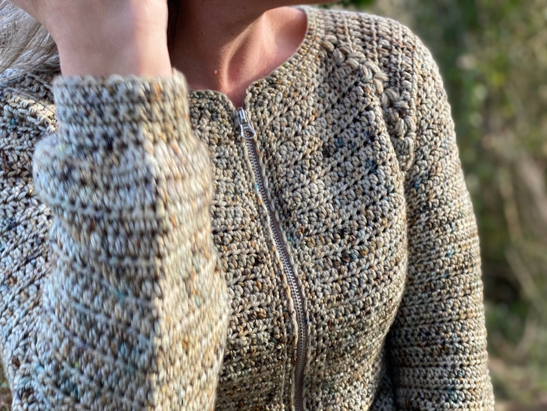 May include: A close-up of a beige crocheted cardigan with a zip fastening. The cardigan features a textured stitch pattern and a braided detail on the shoulder.