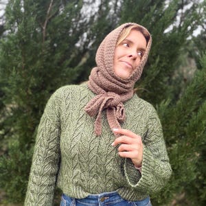 May include: A woman wearing a green knit sweater, blue jeans, and a brown crocheted hood and scarf. The sweater has a textured pattern. The scarf is tied in a bow.