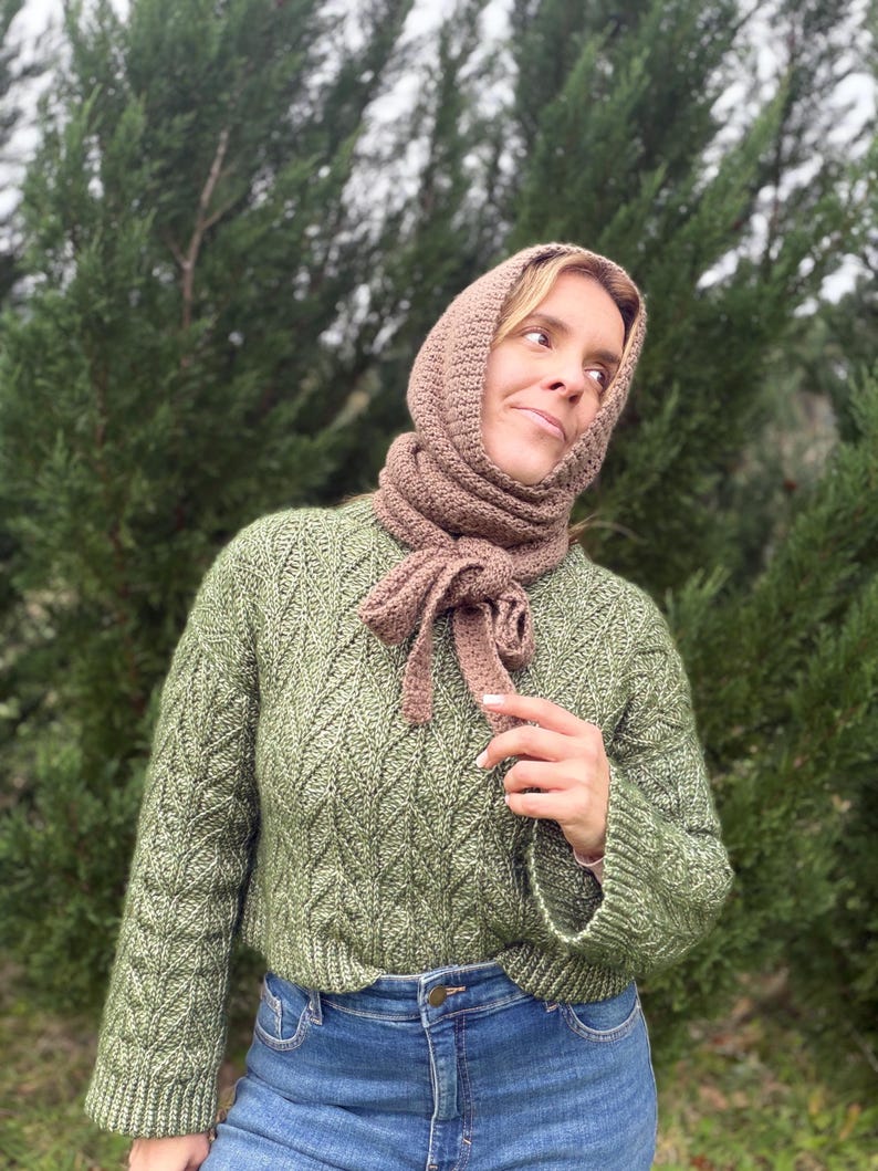 May include: A woman wearing a green knit sweater, blue jeans, and a brown crocheted hood and scarf. The sweater has a textured pattern. The scarf is tied in a bow.