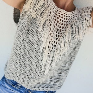 Puede incluir: Un top de crochet gris con un ribete y flecos de crochet blanco. El top es sin mangas y tiene un escote redondo.