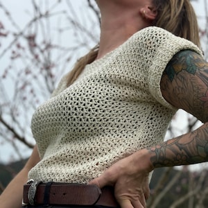 Puede incluir: Top de crochet color crema con tejido abierto y mangas cortas. El top tiene un aspecto texturizado y un ajuste relajado. Se ve un cinturón de cuero marrón con una hebilla plateada. La imagen está tomada al aire libre.