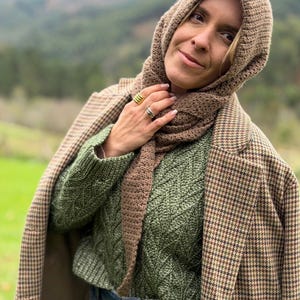 May include: A woman wearing a brown knitted hood and scarf set, paired with a green cable-knit sweater and a brown and black houndstooth blazer. The outfit is completed with blue jeans.