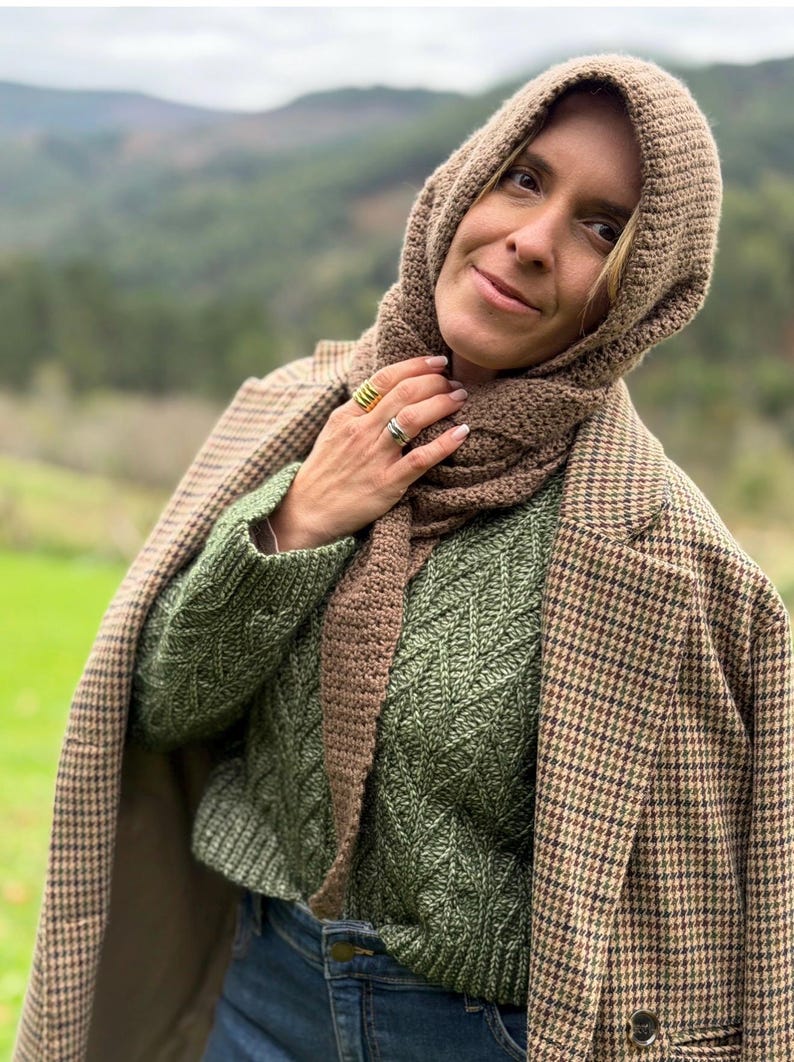 May include: A woman wearing a brown knitted hood and scarf set, paired with a green cable-knit sweater and a brown and black houndstooth blazer. The outfit is completed with blue jeans.
