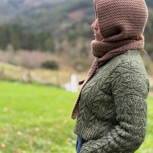 May include: A brown crocheted hood and scarf set paired with a green cable-knit sweater. The hood covers the head and neck, with the scarf draped around the neck. The sweater has a textured pattern. The background features a green field and mountains.