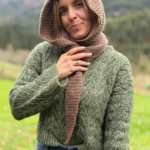May include: A woman wearing a brown crocheted hood and scarf set, paired with a green knit sweater and blue jeans. The hood and scarf are a solid brown color, while the sweater has a textured pattern.
