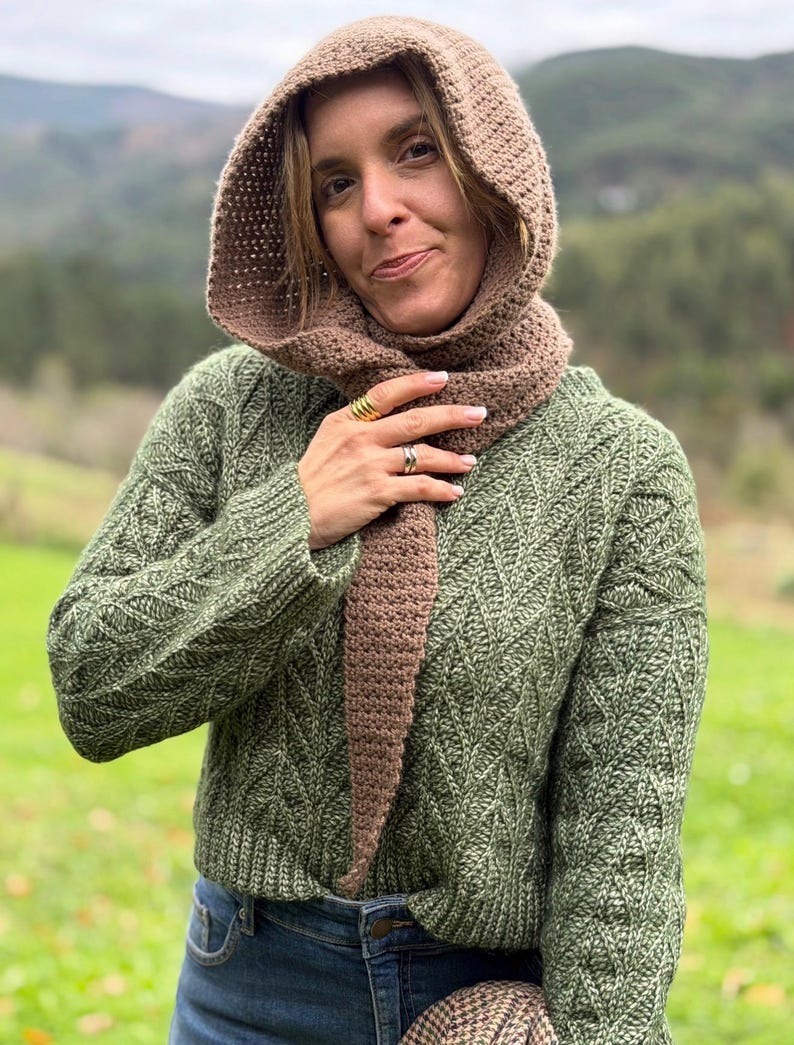 May include: A woman wearing a brown crocheted hood and scarf set, paired with a green knit sweater and blue jeans. The hood and scarf are a solid brown color, while the sweater has a textured pattern.