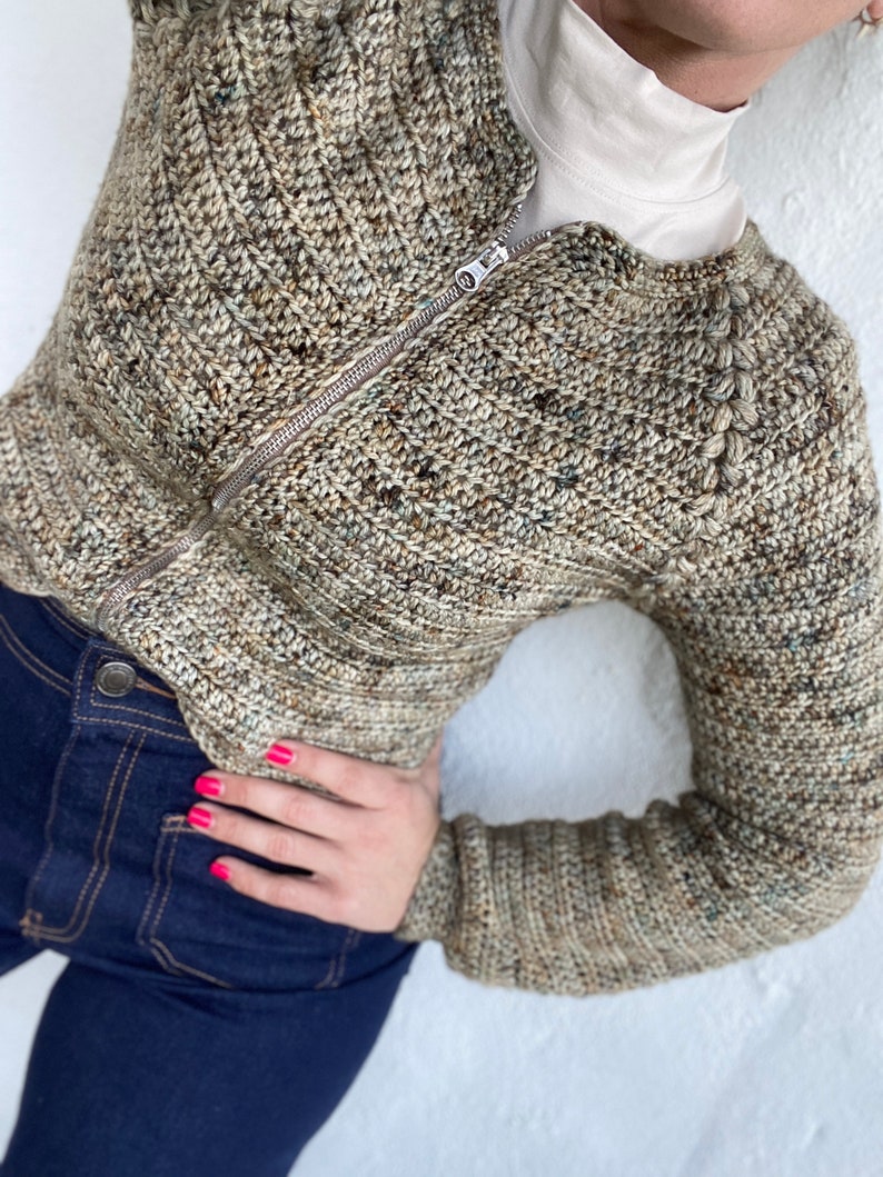 May include: A brown and beige crocheted cardigan with a zip closure. The cardigan has a textured stitch pattern and a high neckline.