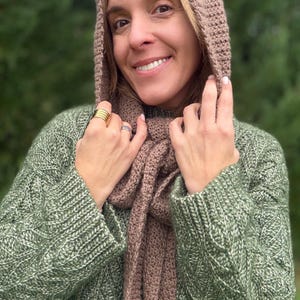May include: A woman wearing a green sweater and a brown knitted hood and scarf. The sweater has a textured pattern. The hood and scarf are a matching color. The woman is smiling and wearing gold and silver rings.