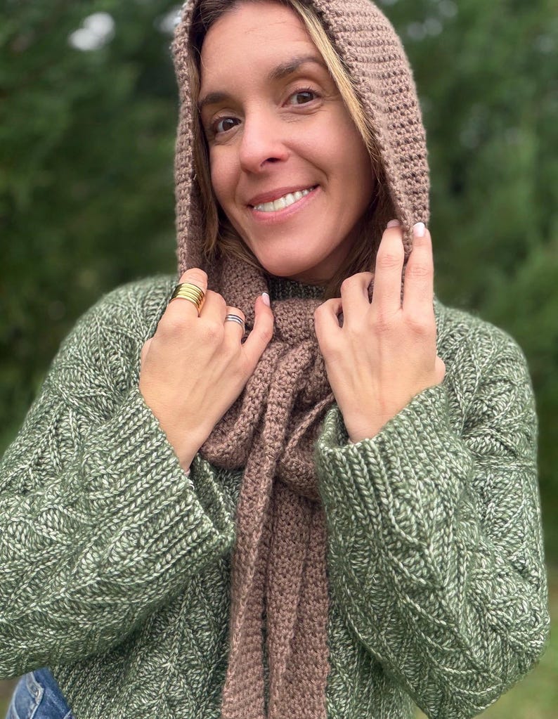 May include: A woman wearing a green sweater and a brown knitted hood and scarf. The sweater has a textured pattern. The hood and scarf are a matching color. The woman is smiling and wearing gold and silver rings.