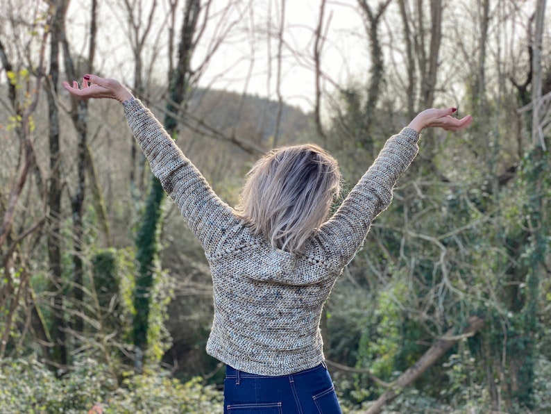 May include: A person wearing a beige and brown crocheted sweater with long sleeves, standing with arms raised in a woodland setting.