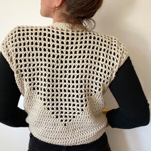 Op de afbeelding: Een beige gebreide vest met een los, opengewerkt patroon. Het vest is mouwloos en heeft een V-hals.