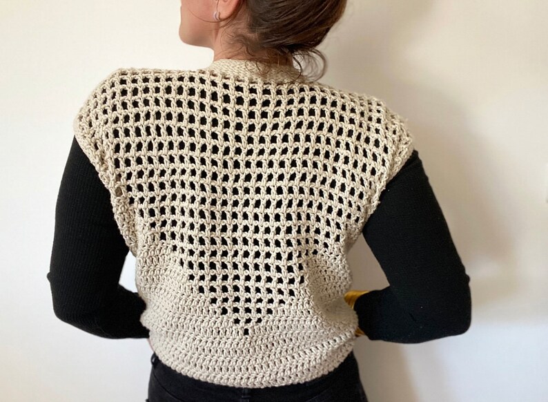 Op de afbeelding: Een beige gebreide vest met een los, opengewerkt patroon. Het vest is mouwloos en heeft een V-hals.