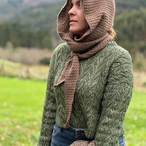 May include: A woman wearing a brown crocheted hood and scarf set. The hood is connected to a scarf that drapes down the front. She is also wearing a green sweater and blue jeans. The background features a green field and a mountain range.