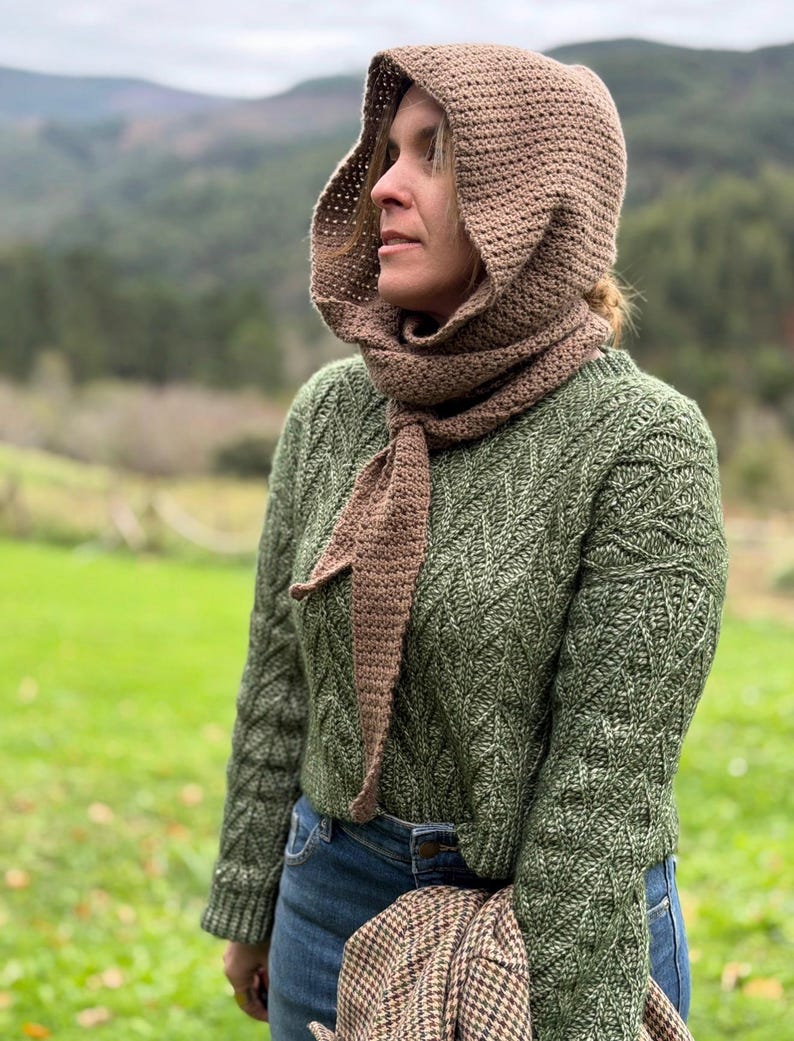 May include: A woman wearing a brown crocheted hood and scarf set. The hood is connected to a scarf that drapes down the front. She is also wearing a green sweater and blue jeans. The background features a green field and a mountain range.