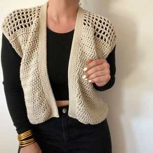 Op de afbeelding: Een beige gebreide vest met een open voorkant en zonder mouwen. De vest is gemaakt met een delicaat open steekpatroon.