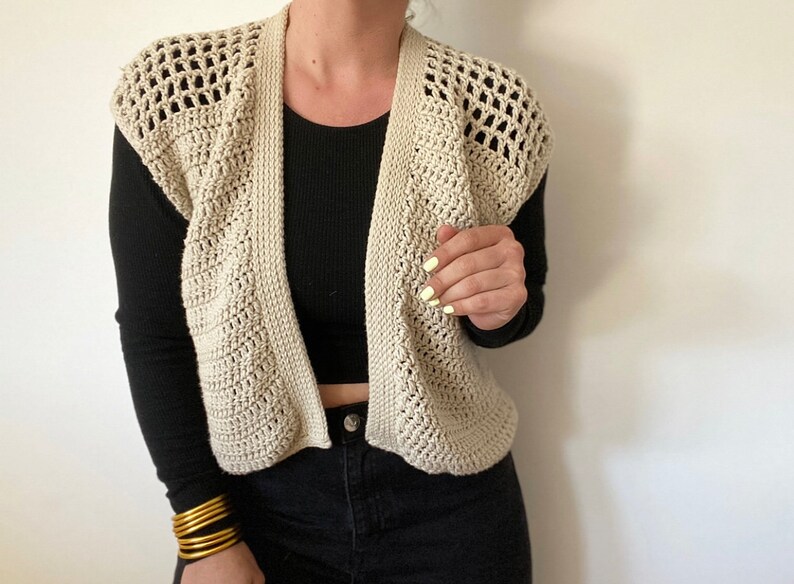 Op de afbeelding: Een beige gebreide vest met een open voorkant en zonder mouwen. De vest is gemaakt met een delicaat open steekpatroon.