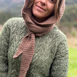 May include: A brown crocheted hood and scarf set. The hood is connected to a long scarf that is tied in a knot. The person is wearing a green sweater with a diamond pattern. The background is a blurred view of a mountain.