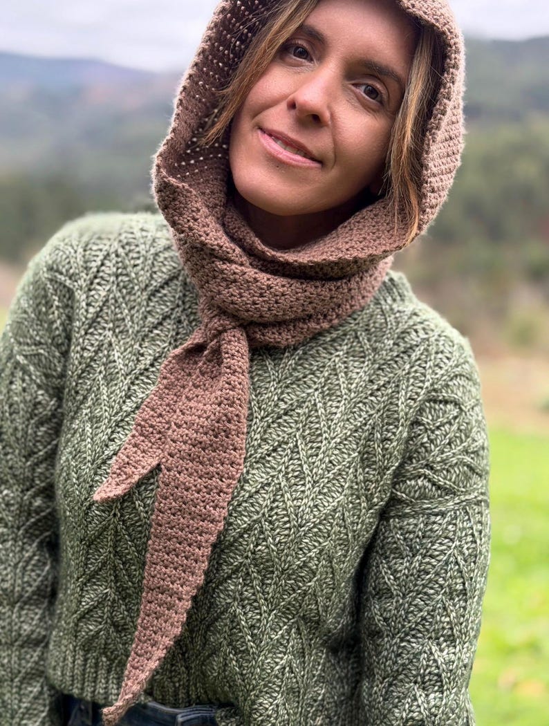 May include: A brown crocheted hood and scarf set. The hood is connected to a long scarf that is tied in a knot. The person is wearing a green sweater with a diamond pattern. The background is a blurred view of a mountain.