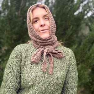 May include: A crocheted hood and scarf set in a muted brown color. The scarf ties in a bow at the neck. The person in the image is wearing a green sweater with a textured pattern. The background is a blurred view of green trees.