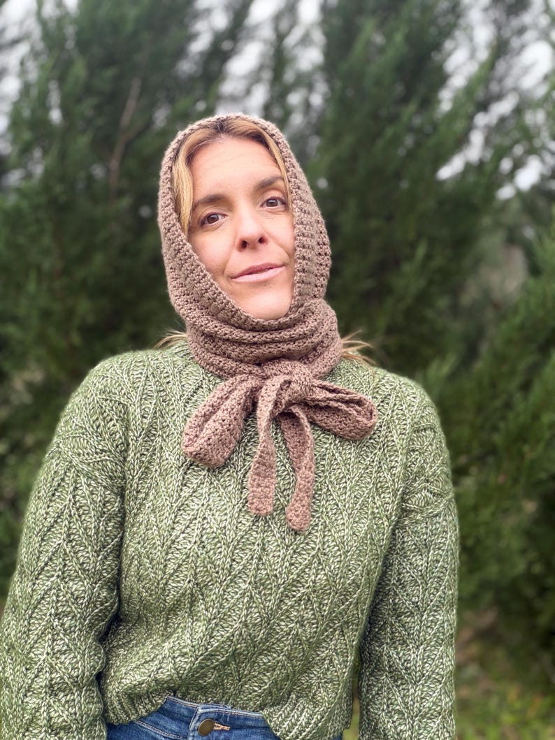 May include: A crocheted hood and scarf set in a muted brown color. The scarf ties in a bow at the neck. The person in the image is wearing a green sweater with a textured pattern. The background is a blurred view of green trees.