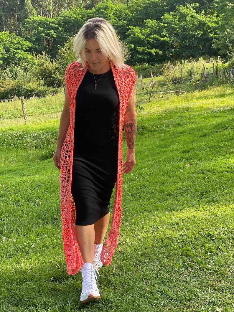 Pu&ograve; includere: Una donna indossa un cardigan di uncinetto corallo sopra un vestito nero e scarpe da ginnastica bianche. Sta camminando in un campo erboso.