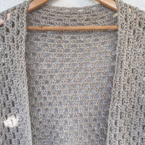 Puede incluir: Un cárdigan de ganchillo beige claro de tejido abierto colgado de una percha de madera. El cárdigan tiene un diseño suelto y aireado con un patrón rectangular. El color neutro y el tejido abierto sugieren una prenda ligera.