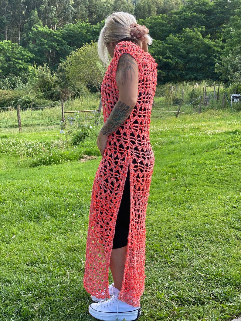 Pu&ograve; includere: Un lungo abito in crochet color corallo con uno spacco laterale. L'abito &egrave; senza maniche e ha una vestibilit&agrave; ampia e fluida. La modella indossa l'abito con leggings neri e sneakers bianche.