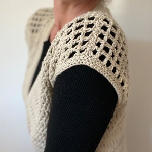 Op de afbeelding: Een beige gehaakte vest met een kantpatroon op de schouders. Het vest wordt gedragen over een zwart longsleeve shirt.