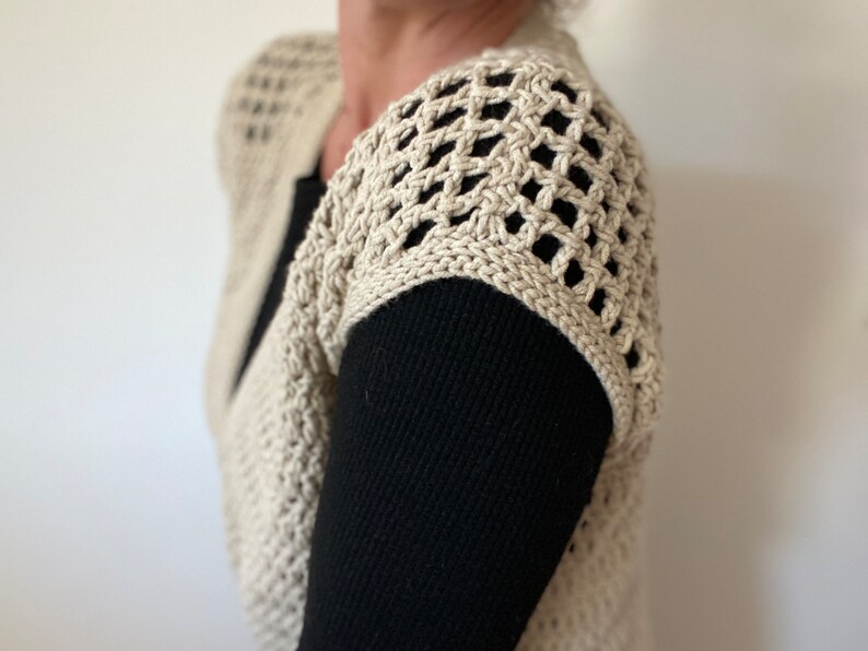 Op de afbeelding: Een beige gehaakte vest met een kantpatroon op de schouders. Het vest wordt gedragen over een zwart longsleeve shirt.